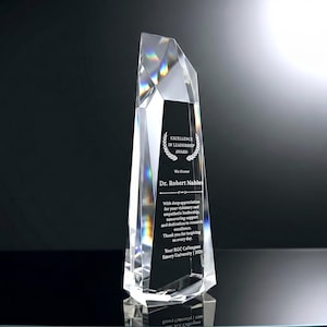 May include: A clear, multifaceted crystal award trophy with a tapered, geometric design. The front features a black panel engraved with the text Excellence in Leadership Award, Dr. Robert Nables, and a message of appreciation from RGC Colleagues.