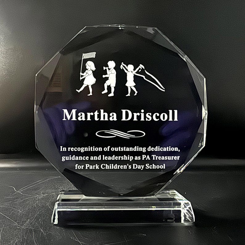 Puede incluir: Un premio de cristal claro y facetado con una base rectangular. El premio presenta el nombre "Martha Driscoll" y una inscripci&oacute;n que reconoce la dedicaci&oacute;n excepcional. Un gr&aacute;fico de ni&ntilde;os est&aacute; grabado en la parte superior.