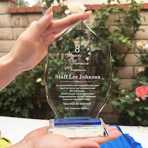 Puede incluir: Un premio de cristal transparente y facetado con una base azul. El premio dice "8 Years of Service" y se presenta a Staff Lee Johnson. El texto incluye agradecimiento por el servicio y felicitaciones por la jubilación. El premio se sostiene en las manos.