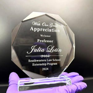 Puede incluir: Un premio de cristal transparente y facetado con el texto "With Our Great Appreciation" y "Professor Julia Lotin". El premio tambi&eacute;n incluye el texto "Southwestern Law School Externship Program 2024". El premio lo sostiene una persona con un guante morado.