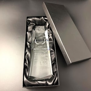 May include: A clear, faceted glass award trophy resting inside a black rectangular gift box lined with black satin fabric. The award features the engraved text Excellence in Leadership Award, We Honor Dr. Robert Nables, and additional appreciation text.