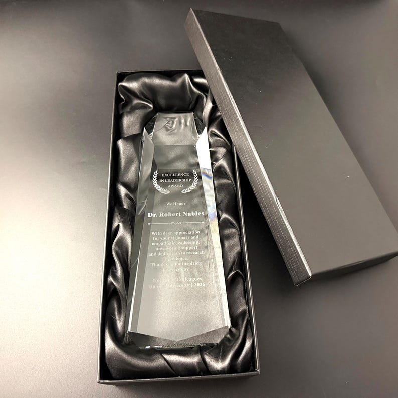 May include: A clear, faceted glass award trophy resting inside a black rectangular gift box lined with black satin fabric. The award features the engraved text Excellence in Leadership Award, We Honor Dr. Robert Nables, and additional appreciation text.