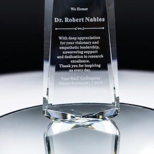 May include: A clear glass award trophy with beveled edges, featuring an engraved inscription honouring Dr. Robert Nables for visionary leadership and research excellence, presented by RGC Colleagues at Emory University in the year 2026.