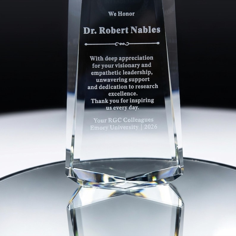 May include: A clear glass award trophy with beveled edges, featuring an engraved inscription honouring Dr. Robert Nables for visionary leadership and research excellence, presented by RGC Colleagues at Emory University in the year 2026.