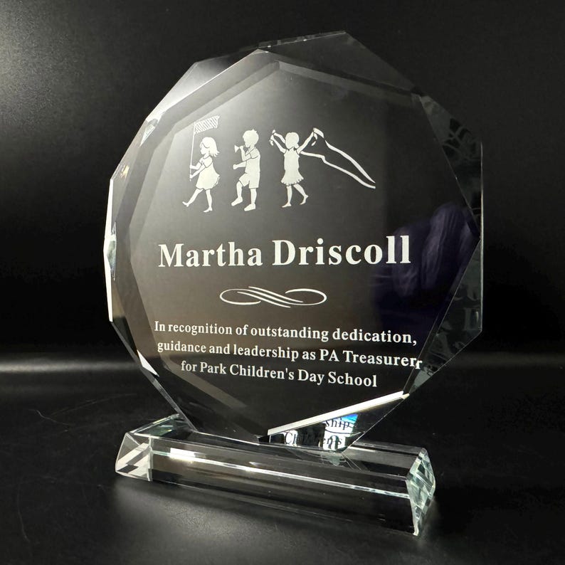 Puede incluir: Un premio de cristal transparente y facetado con una base rectangular. El premio presenta el nombre "Martha Driscoll" y una inscripci&oacute;n de reconocimiento. Un grabado de ni&ntilde;os adorna el cristal.