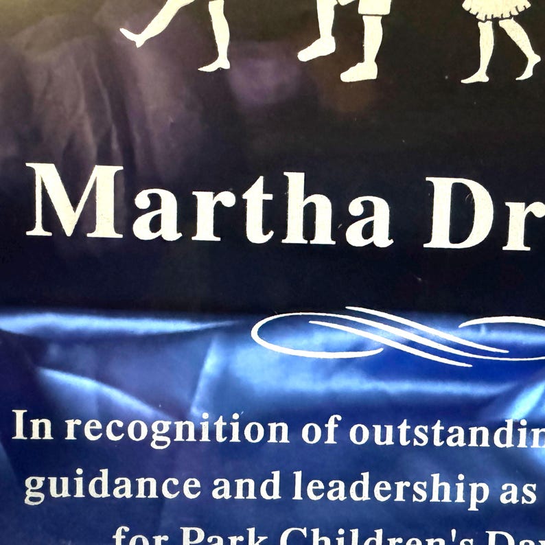Puede incluir: Placa azul marino con texto blanco "Martha Dr". Debajo, el texto contin&uacute;a: "En reconocimiento a la destacada orientaci&oacute;n y liderazgo como para el D&iacute;a de los Ni&ntilde;os del Parque". Siluetas de ni&ntilde;os en la parte superior.