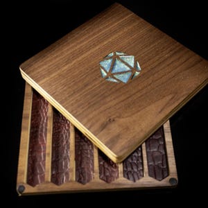 May include: A wooden box with a hinged lid, featuring a geometric design inlaid with blue and silver accents. The box is open, revealing several textured, dark brown leather strips. The box is set against a black background.