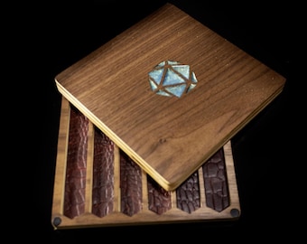 Puede incluir: Una caja de madera con tapa con bisagras, con un diseño geométrico con incrustaciones de detalles azules y plateados. La caja está abierta, revelando varias tiras de cuero marrón oscuro texturizado. La caja está sobre un fondo negro.