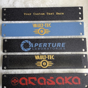 Op de afbeelding: Een verzameling rechthoekige metalen platen in zwart en verschillende kleuren, met logo's en tekst. De platen tonen "Your Custom Text Here", "VAULT-TEC", "APERTURE LABORATORIES" en "ARASAKA" in verschillende kleuren.