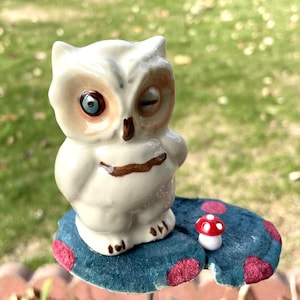 Puede incluir: Un búho de cerámica blanca con un ojo azul y un ojo cerrado, posado sobre una base en forma de hongo azul y rojo.