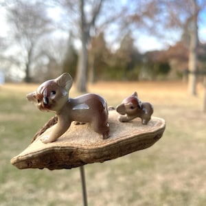 May include: Two small ceramic koala figurines on a wooden base. The larger koala is brown and has a light brown belly. The smaller koala is brown and has a light brown belly. The base is a light brown color and has a rough texture.