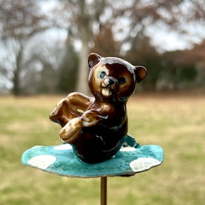 May include: A brown ceramic bear figurine with blue eyes, sitting on a teal and white polka dot mushroom. The mushroom is attached to a gold metal stick.