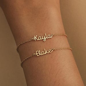 May include: Two delicate gold bracelets, each with a name in cursive script: "Kayla" and "Blake." The bracelets are worn on a wrist, showcasing the personalized jewelry. The background is a neutral beige.