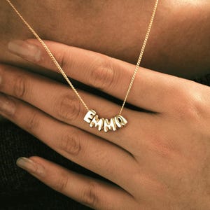 Puede incluir: Un collar dorado con el nombre "EMMA" escrito con cuentas de letras individuales. El collar se lleva alrededor del cuello. El collar está sujeto por una mano con uñas largas.