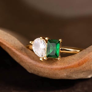 May include: A gold ring featuring a pear-shaped white gemstone and a rectangular emerald-cut green gemstone. The ring is set on a gold band and rests on a brown, textured surface.
