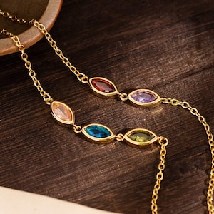 May include: Two gold necklaces, each with three oval-cut gemstones in various colours. One necklace has red, purple, and yellow-green stones. The other has peach, blue, and yellow-green stones. The necklaces are on a dark wooden surface.