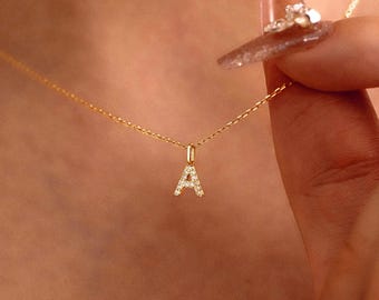 May include: A delicate gold chain necklace featuring a small, capital letter A pendant. The letter A is encrusted with tiny, clear, sparkling stones that catch the light. The necklace rests against the skin, showcasing a simple and refined design.