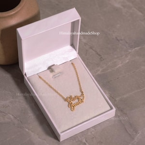 May include: A gold-toned necklace with a cursive nameplate reading Olivia Brooks and a small heart charm, displayed inside an open white rectangular jewelry box. The text HimalayahandmadeShop is visible on the box lid and the interior lining.