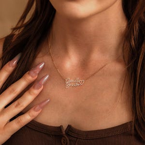May include: A gold-toned metal necklace featuring a delicate chain and a personalized pendant with the names Camille and Braxton written in a cursive script font, accompanied by a small open heart outline attached to the end of the names.