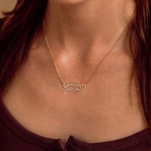 May include: A gold-toned metal necklace featuring a delicate chain and a personalized nameplate pendant. The pendant displays the script text Camille Braxton with a small open heart symbol integrated into the design at the end of the name.