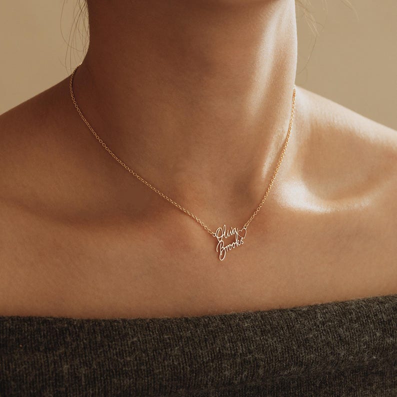 May include: A gold-toned metal necklace featuring a delicate chain and a personalized pendant with the name Olivia Brooks written in a cursive script font, accompanied by a small open heart outline attached to the end of the name.