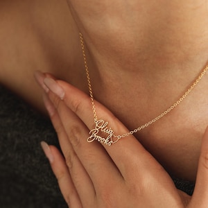 May include: A gold-toned metal necklace featuring a delicate chain and a personalized pendant. The pendant displays the names Olivia and Brooks in a cursive script font, positioned one above the other, with a small heart outline to the right.