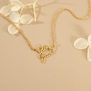 May include: A gold-toned metal necklace featuring a delicate chain and a custom nameplate pendant. The pendant displays the names Oluw and Brooks in a cursive script font, positioned one above the other, with a small open heart outline on the right.