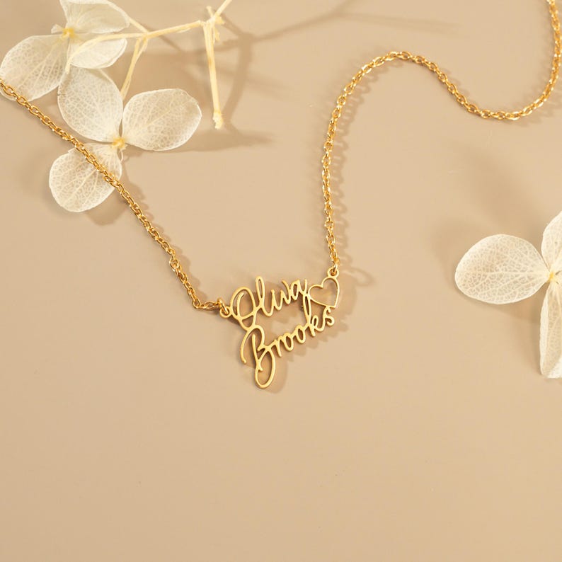 May include: A gold-toned metal necklace featuring a delicate chain and a custom nameplate pendant. The pendant displays the names Oluw and Brooks in a cursive script font, positioned one above the other, with a small open heart outline on the right.