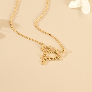 May include: A gold-toned metal necklace featuring a delicate chain and a personalized pendant with the names Oluwa and Brooks written in a cursive script font, accompanied by a small open heart shape integrated into the right side of the design.