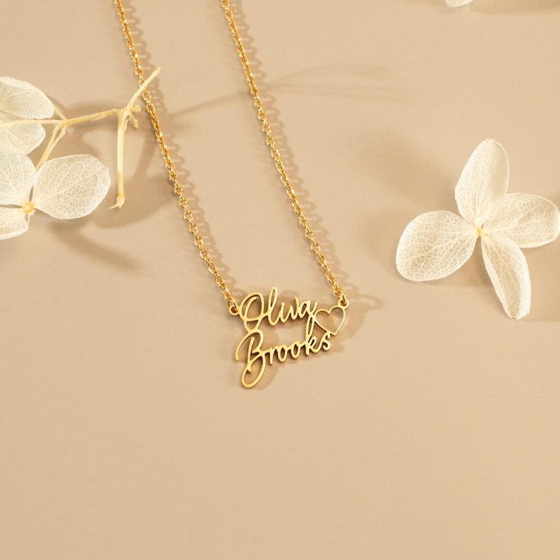 May include: A gold-toned necklace featuring a delicate chain and a personalized pendant with the script text "Oliva Brooks" arranged in two lines, accompanied by a small open heart outline integrated into the right side of the name design.