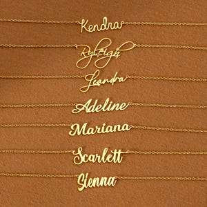Könnte beinhalten: Goldfarbene Halsketten mit personalisierten Namensanhängern. Die Namen Kendra, Ryleigh, Leandra, Adeline, Mariana, Scarlett und Sienna sind in einer Schreibschrift dargestellt. Die Halsketten befinden sich auf braunem Hintergrund.