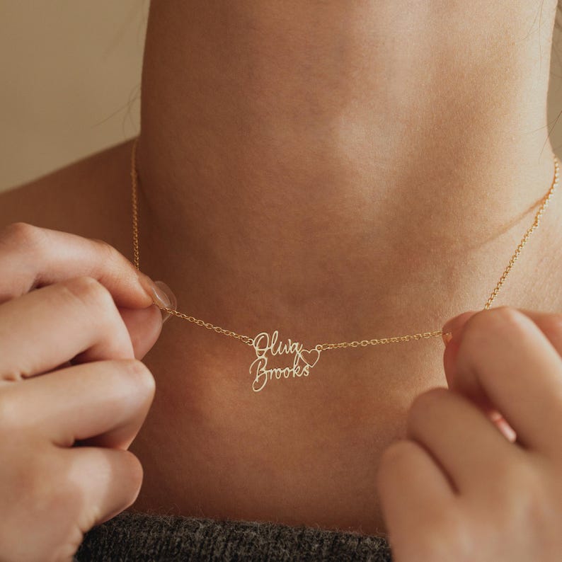 May include: A gold-toned necklace featuring a delicate chain and a personalized pendant with the names "Olwa" and "Brooks" written in a cursive script font, accompanied by a small, open-lined heart symbol integrated into the design.