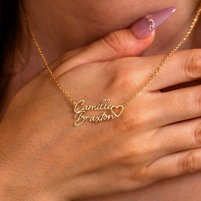 May include: A gold-toned metal necklace featuring a delicate chain and a personalized nameplate pendant. The pendant displays the names Camille and Braxton in a cursive script font, with a small heart outline integrated into the design.