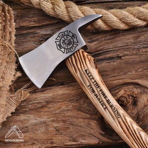 Pode incluir: Um machado de bombeiro em metal prateado com cabo de madeira. A cabeça do machado apresenta um emblema Fire Dept gravado a preto. O cabo tem gravado o texto KEEP THE FIRE BURNING FOR JUSTICE. O machado está sobre um fundo de madeira rústica.