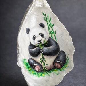 May include: Hand-painted oyster shell art featuring a panda bear eating bamboo. The panda is black and white, with green bamboo stalks and leaves. The shell is white with a textured surface. The artwork is set against a dark gray background.