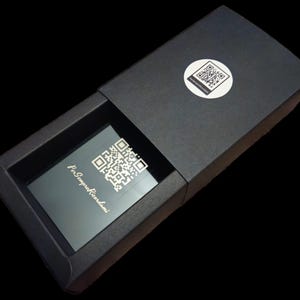 May include: A black box with a silver inscription that reads "Per Sempre Ricordi" and a QR code. The box is open and the inside is lined with black velvet.