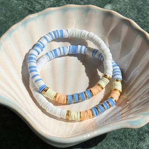 May include: Two beaded bracelets in a seashell-shaped dish. One bracelet features white and light blue beads, the other has white, light blue, peach, and yellow beads. Gold-coloured spacers separate the colours. The dish is pale pink and blue.
