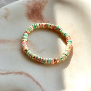 May include: A pastel-coloured bracelet with flat, circular beads in pink, green, and cream, separated by gold-toned spacers. The bracelet is round and likely made from lightweight materials, suitable for daily wear. The circumference is approximately 18 cm.
