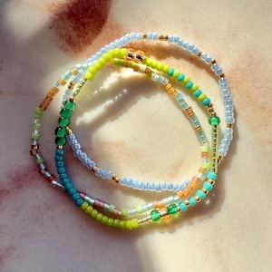 May include: Three beaded bracelets in shades of blue, green, and gold. The bracelets are made of small, colourful beads strung together. The bracelets are stacked, creating a layered effect. The background is a light, textured surface.