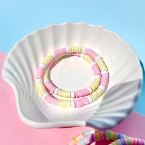 Könnte beinhalten: Zwei Armbänder aus bunten Perlen in Rosa, Gelb und Weiß. Die Armbänder werden in einer weißen Muschelform präsentiert.