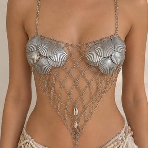 Può includere: Un top a catena color argento composto da una rete di maglie metalliche. Il capo è decorato con diverse conchiglie di capasanta metalliche sulla zona del petto e tre piccole conchiglie di ciprea bianche appese verticalmente al centro.