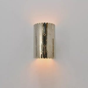 May include: A cylindrical, silver-colored wall sconce with a perforated starburst pattern. The sconce is illuminated from within, casting a warm glow. The fixture is mounted on a white wall, creating a modern lighting effect.