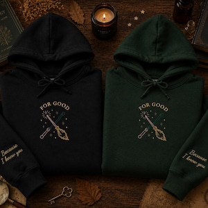 May include: A black hoodie and a dark green hoodie, each featuring a hood with drawstrings. The chest displays an embroidered graphic of a wand and a broomstick crossed with the text FOR GOOD. The sleeve is embroidered with Because I knew you.