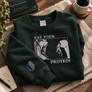 May include: A dark forest green crewneck sweatshirt featuring a white embroidered graphic of a wide-mouthed creature and a hand holding a piece of meat, with the text EAT YOUR PROTEIN. A small blue and white shield emblem is on the left sleeve.