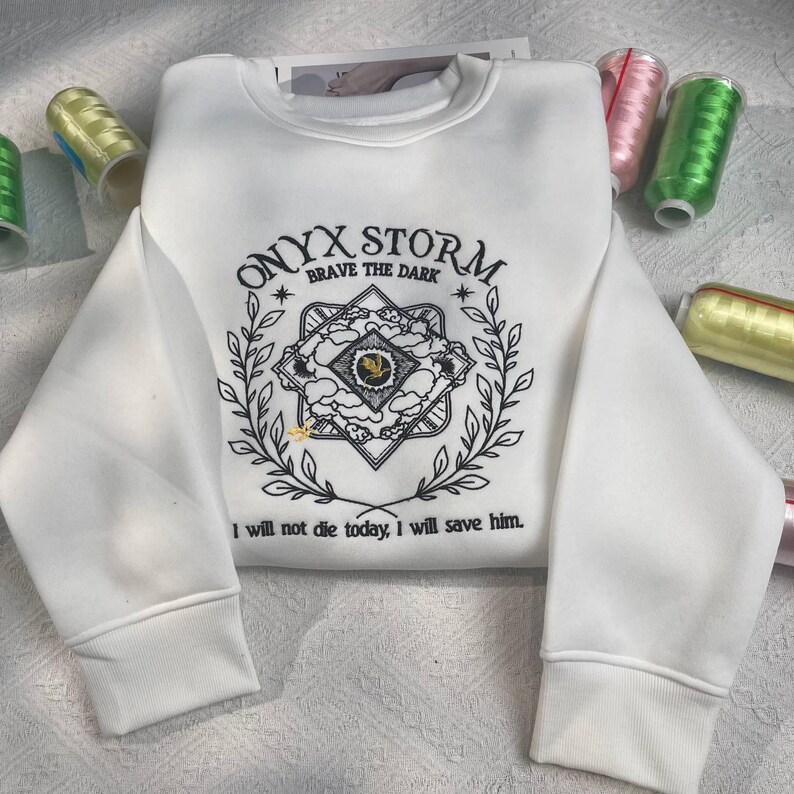 Może przedstawiać: Biała bluza z czarnym haftowanym wzorem geometrycznym z tekstem "ONYX STORM BRAVE THE DARK" i tekstem "Nie umrę dzisiaj, uratuję go."