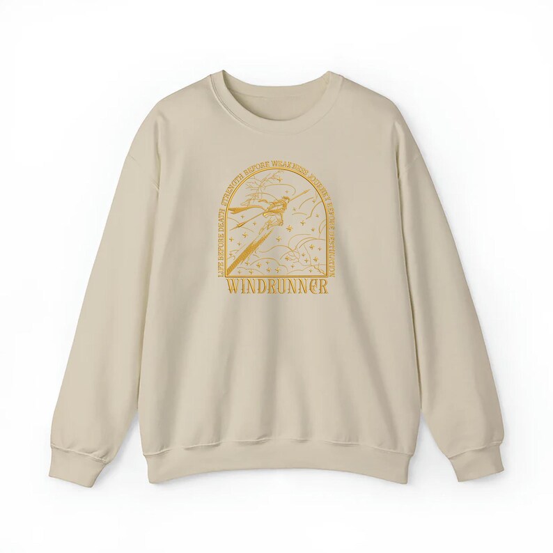 Puede incluir: Sudadera beige con cuello redondo. La parte delantera presenta una ilustraci&oacute;n dorada de una figura con una espada, rodeada de texto que incluye "WINDRUNNER". El dise&ntilde;o est&aacute; centrado en el pecho.