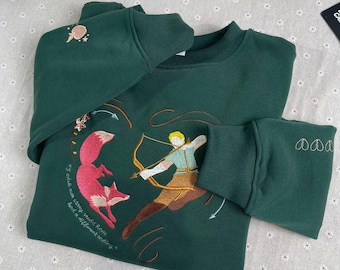 May include: A dark green sweatshirt with an embroidered design of a fox and a person with a bow and arrow. The text "my neck was strong with roses, had to differentiate and it"