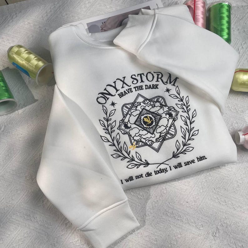 Może przedstawiać: Biała bluza z czarnym haftowanym tekstem, kt&oacute;ry brzmi "ONYX STORM. BRAVE THE DARK" i grafiką gwiazdy z chmurami i liśćmi. Tekst pod grafiką brzmi "Nie umrę dzisiaj, uratuję go."