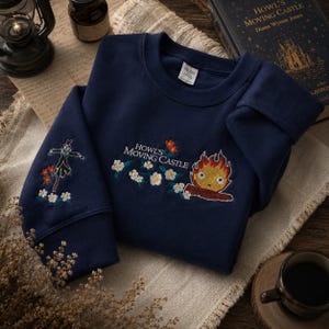 May include: Navy blue sweatshirt with embroidered designs from "Howl's Moving Castle." Features a scarecrow on one sleeve and a fire demon with the text "Howl's Moving Castle" on the chest, surrounded by flowers. The sweatshirt is folded on a neutral-colored surface.