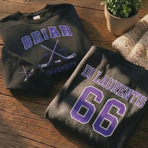 Puede incluir: Dos sudaderas negras de cuello redondo con letras moradas. Una muestra 'BRIAR UNIVERSITY' con palos de hockey cruzados y 'EST 2013'. La otra lleva 'DI LAURENTIS 66' en la espalda. Un pequeño gráfico de palos de hockey cruzados está en la manga.